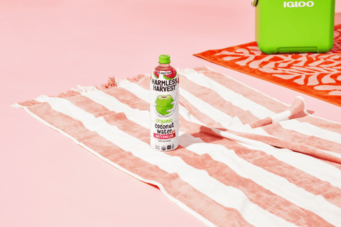 harmless harvest watermelon organic coconut water bottle sitting on a pink and white striped towel on a pink background