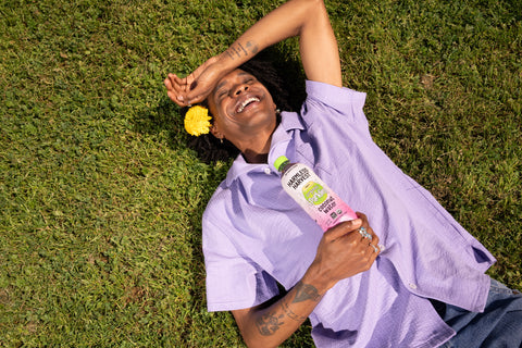 man laying on the grass in purple shirt holding harmless harvest sparkling coconut water bottle