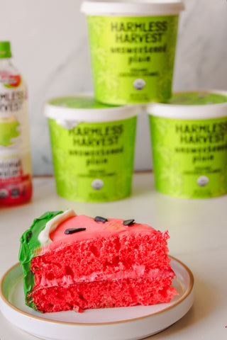 a slice of watermelon cake plated in front of a stack of harmless harvest dairy-free yogurt alternative tubs and a bottle of watermelon organic coconut water